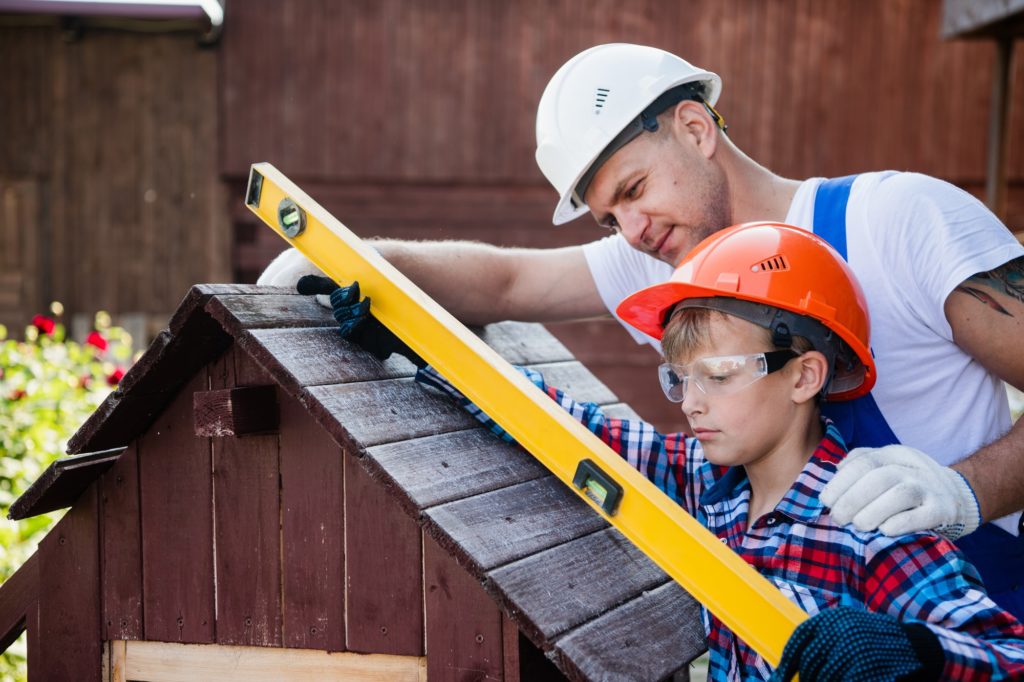 Happy father looking at son measuring wooden plank with building level. Building a dog house at